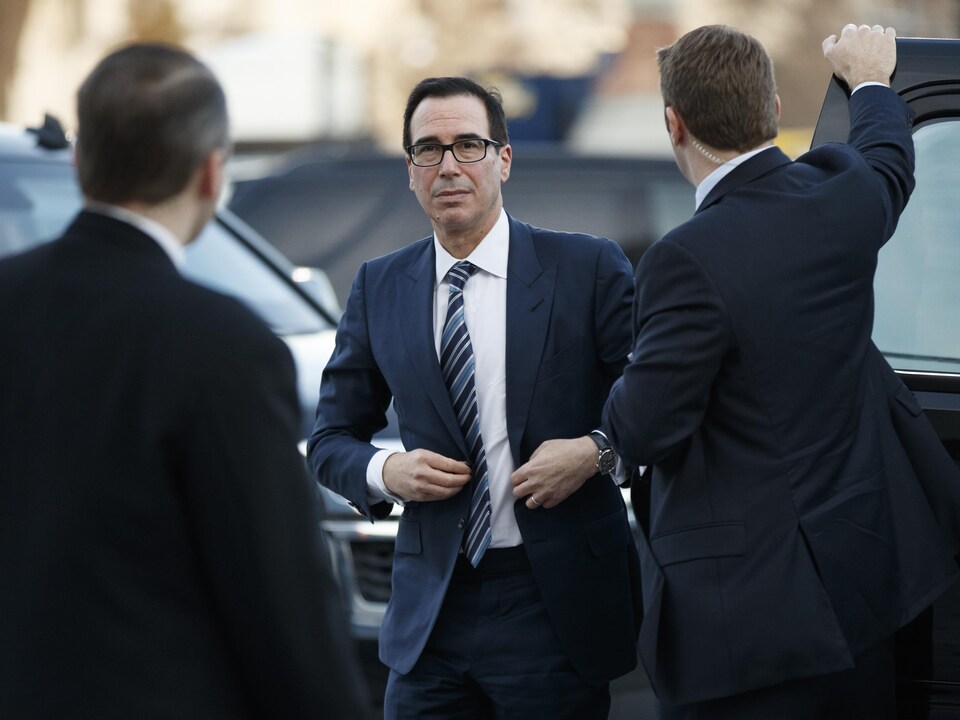 Un homme portant des lunettes et un complet avec une cravate sort d'un véhicule noir entouré de deux agents de sécurit