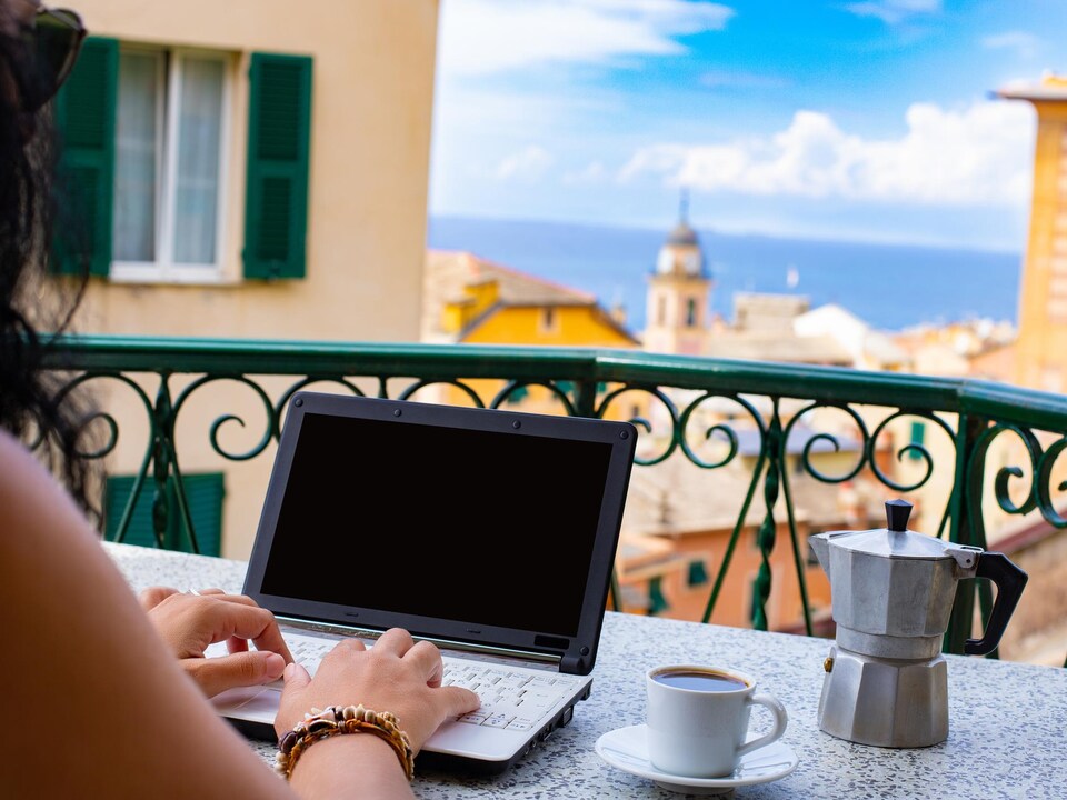 Une femme travaille sur son portable sur un balcon avec vue sur la mer et les toits de la ville.