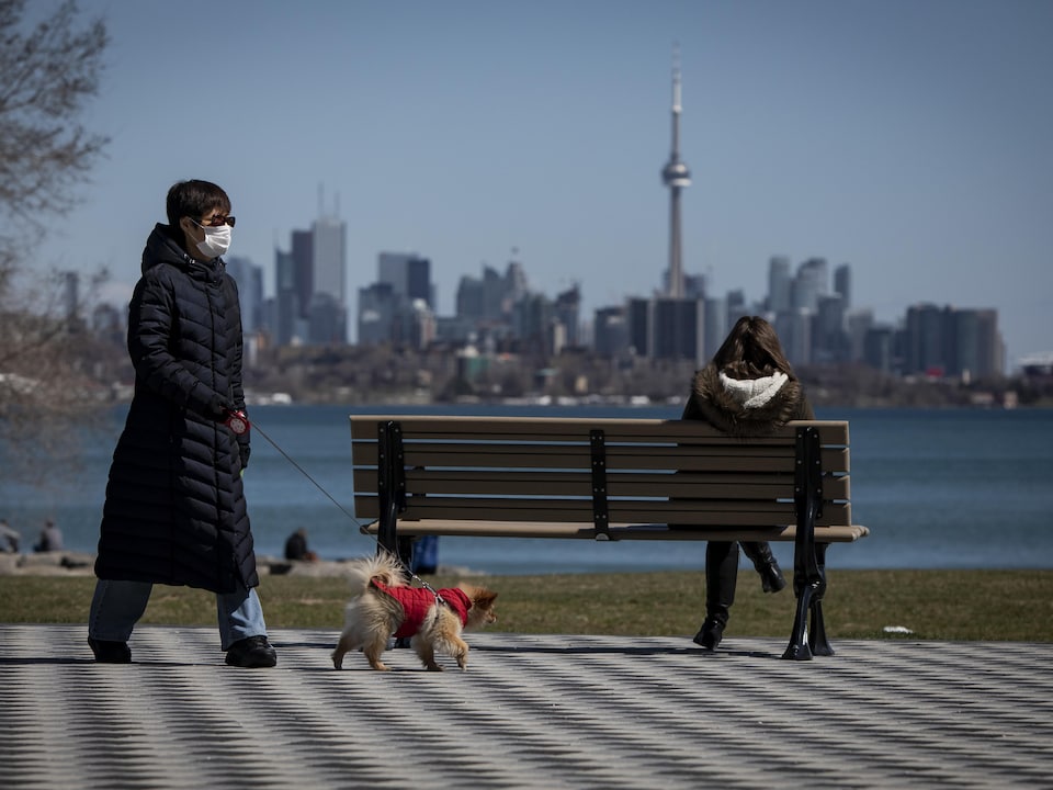 Une femme avec un masque passe à distance d'un banc public où est assise une autre femme qui regarde la ville de Toronto.