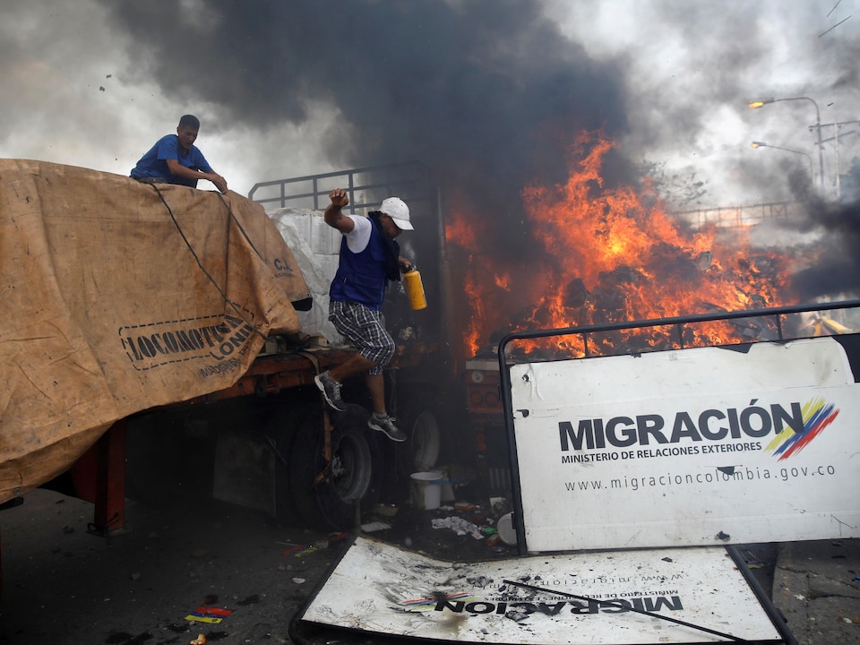 Deux hommes travaillent entre deux camions sur une route. L'un des deux véhicules est en feu.