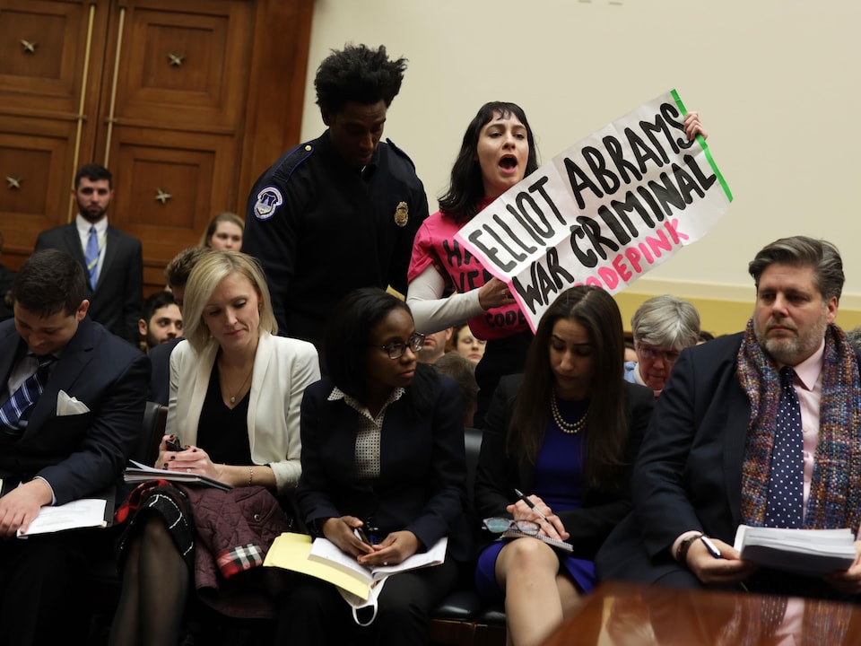 Une femme tient une affiche sur laquelle on peut lire : Elliott Abrams, criminel de guerre. Elle est conduite hors de la salle par un agent de sécurité.