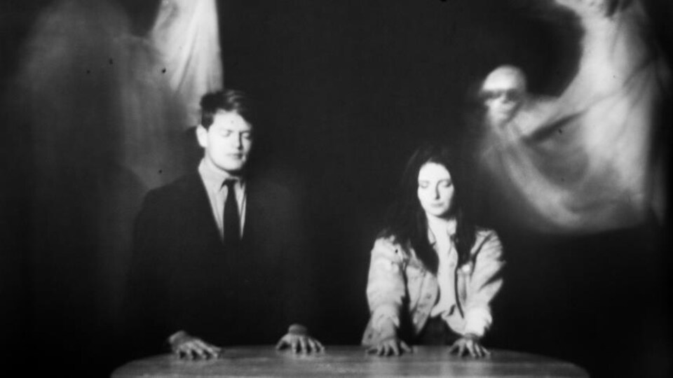 Un homme et une femme assis les yeux fermés à une table. Leurs mains sont posées sur la table. Autour d'eux des fantômes. 