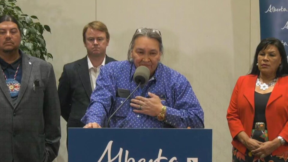 Le chef Douglas Beaverbones se tient debout derrière un podium, la main sur la poitrine. Derrière lui se trouve le ministre de la santé Tyler Shandro.
