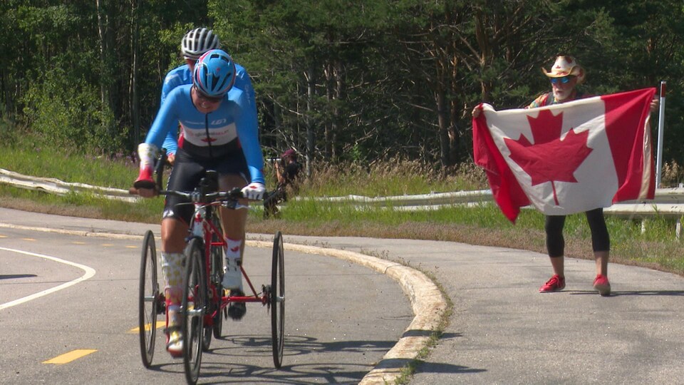 Shelley Gautier est sur un vélo et sur le bord de la route, son conjoint Alan Geer tient un drapeau du Canada. 