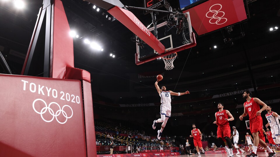 Le basketteur américain s'apprête à déposer le ballon dans le panier à la suite d'un saut. Trois joueurs adverses le regardent avec attention.