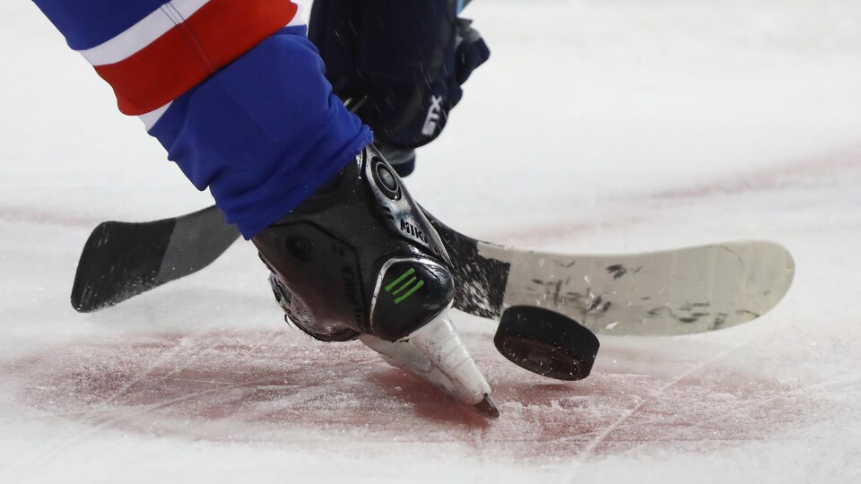 Deux bâtons de hockey se disputent la rondelle entre des patins lors d'un match.