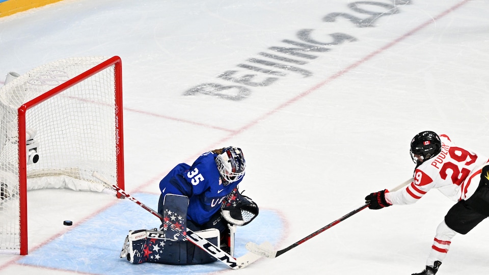 Une joueuse de hockey en blanc et rouge marque un but face à une gardienne en bleu.