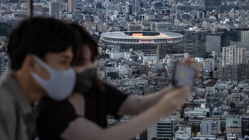 Deux jeunes Japonais prennent un égoportrait avec, en arrière-plan, le stade olympique de Tokyo.