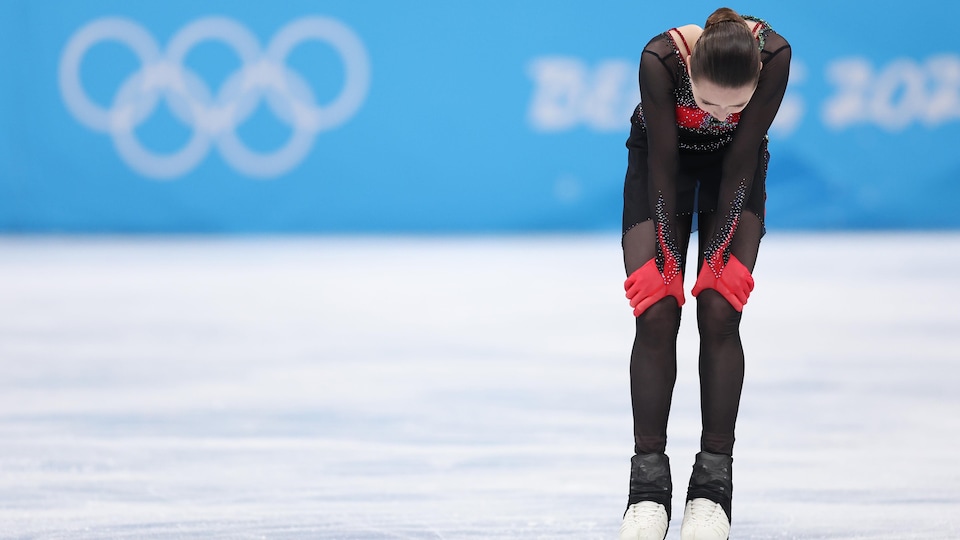 Une patineuse est penchée et se tient les genoux après sa prestation sur la glace. On voit le logo olympique en arrière-plan.