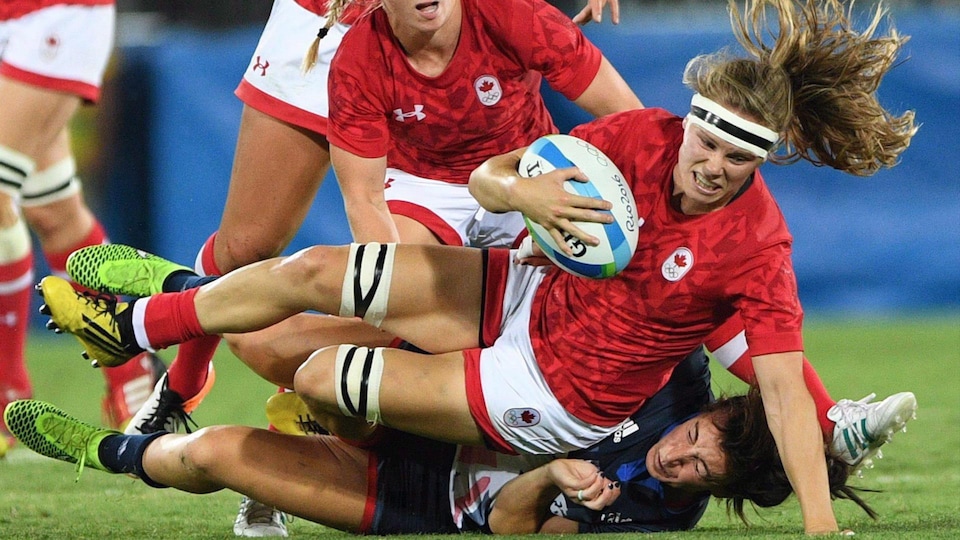 Une joueuse canadienne de rugby aux longs cheveux blonds tient le ballon du bras droit, alors qu'elle tombe sur le terrain.