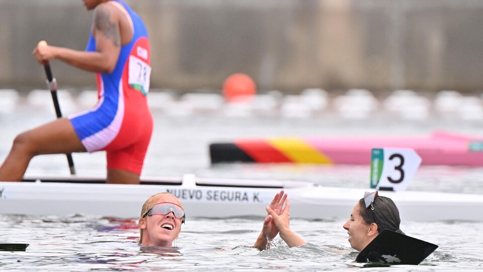 Deux femmes sont dans l'eau. Elles se donnent la main, pendant qu'une autre femme pagaie sur son canot à l'arrière-plan.