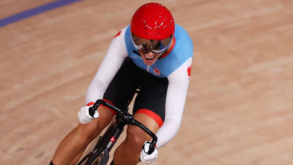 La cycliste, en combinaison aérodynamique blanche et bleue avec un casque rouge, pédale fort sur une piste de cyclisme. 