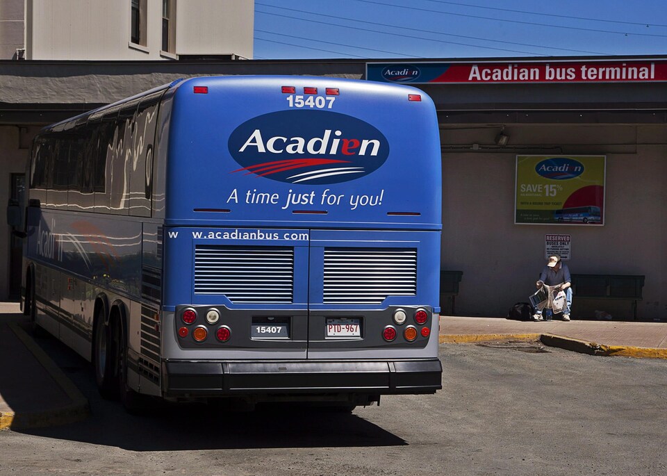 Un autobus bleu d'Acadian Lines est stationné au terminus. Un homme est assis le long du bâtiment, la tête penchée sur un journal.