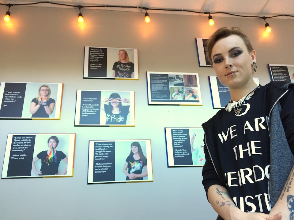 Aédan Charest pose devant un mur arborant des portraits issus de son projet artistique sur la diversité sexuelle.