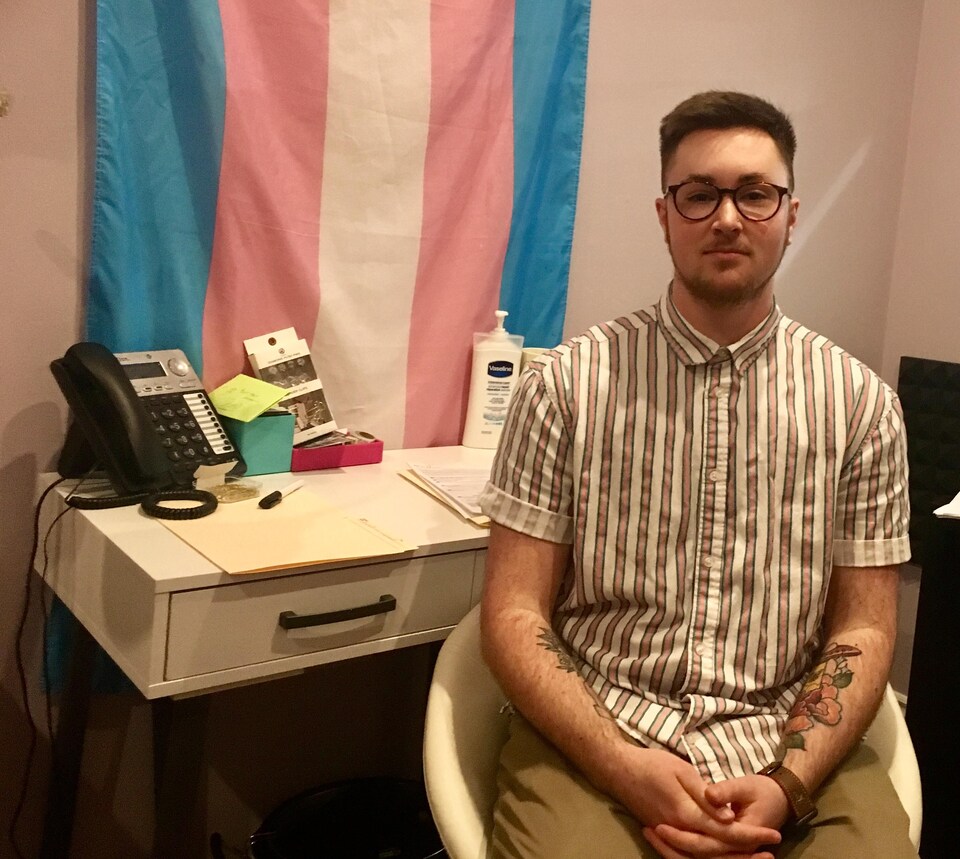 Un homme est assis, il porte une chemine bleue, blanche et rose, à l'effigi des couleurs du drapeau de la fierté trans qui se trouve derrière lui. 