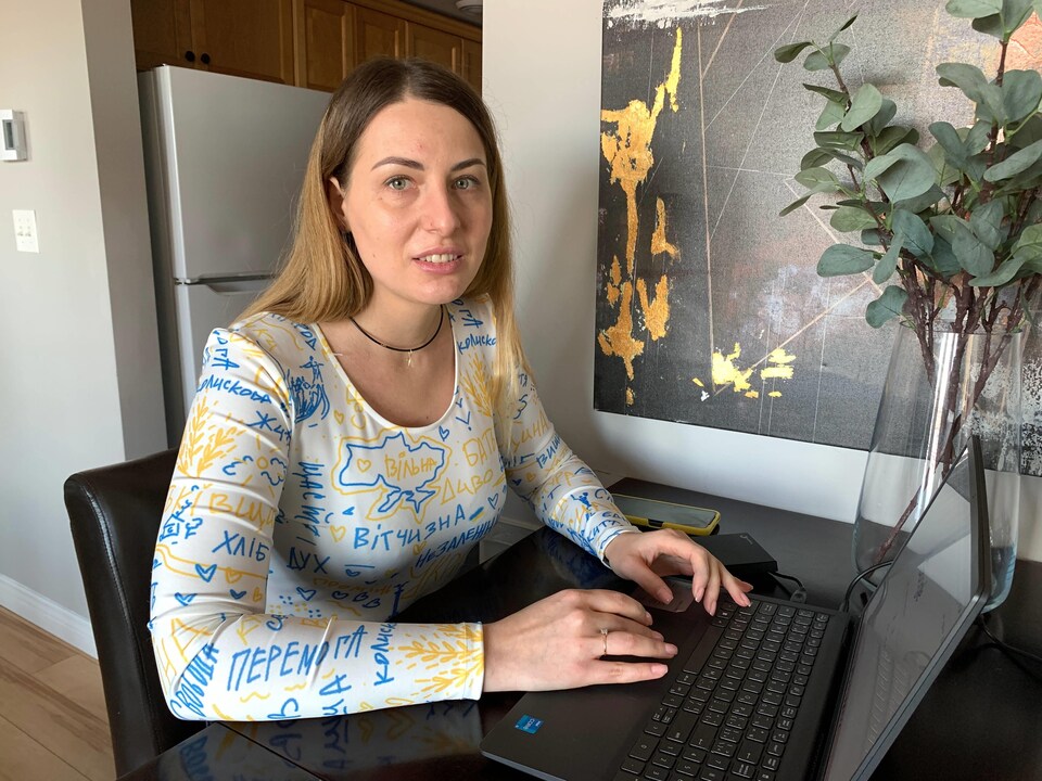 Une femme qui porte une blouse blanche, bleue et jaune aux couleurs du drapeau de l'Ukraine est assise à une table de cuisine. Elle a les deux mains sur le clavier d'un ordinateur portable et lève la tête pour regarder la personne qui la prend en photo.
