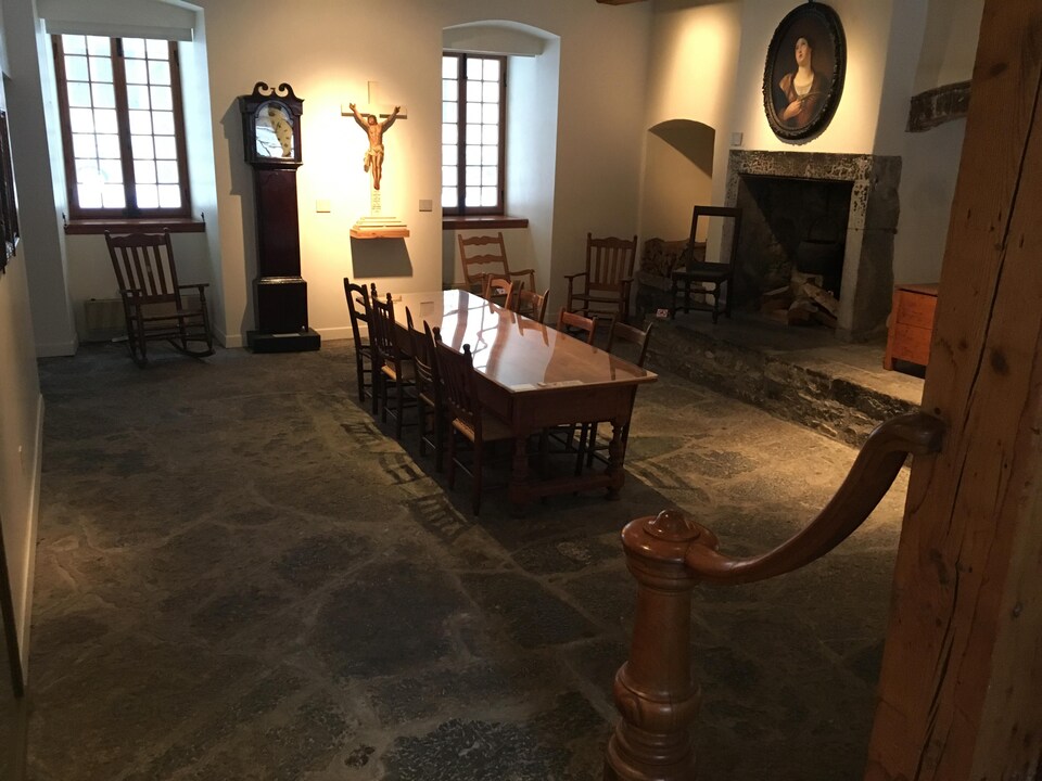 La salle comprends notamment un table, des chaises, un âtre à cheminée et une horloge. Le sol est en pierre et le soleil pénètre dans la salle par deux fenêtres.