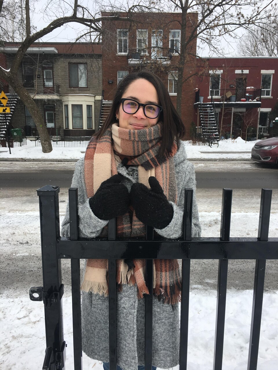 Une jeune femme, portant des lunettes, un manteau et un foulard, est debout derrière une petite clôture dans un parc.