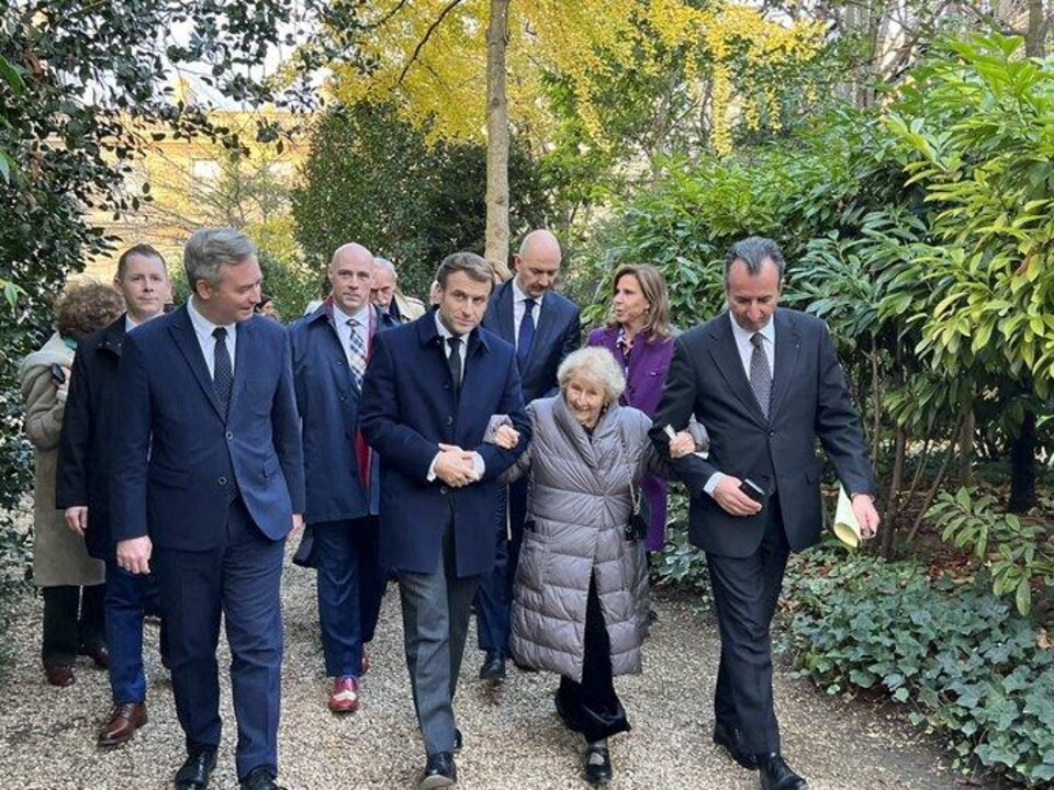 Antonine Maillet donne le bras à Emmanuel Macron et ils marchent au milieu d'un groupe de personnes à l'extérieur.