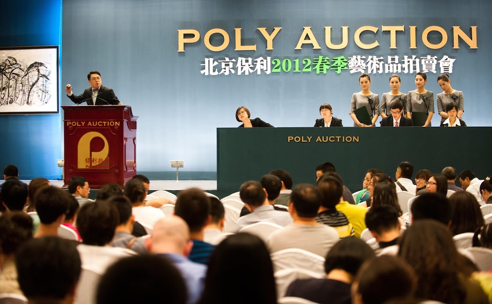 Un homme chinois derrière un lutrin lève son bras droit devant une foule de personne réunies lors de la vente aux enchères d'une oeuvre d'art.