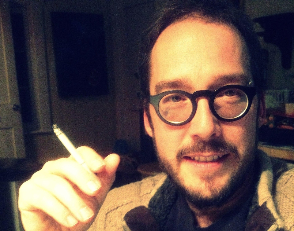 L'écrivain et éditeur Guy D'Amours. Il s'agit d'un bel homme, fin quarantaine qui porte des lunettes et a une cigarette entre les doigts.