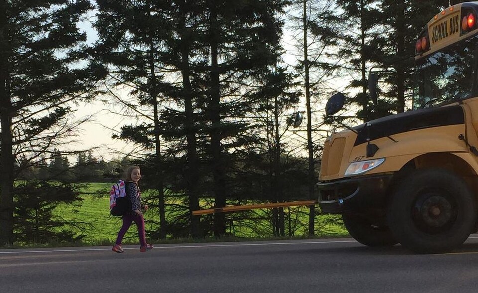 Une enfant souriante traverse une route devant un autobus scolaire dont les feux clignotants sont activés