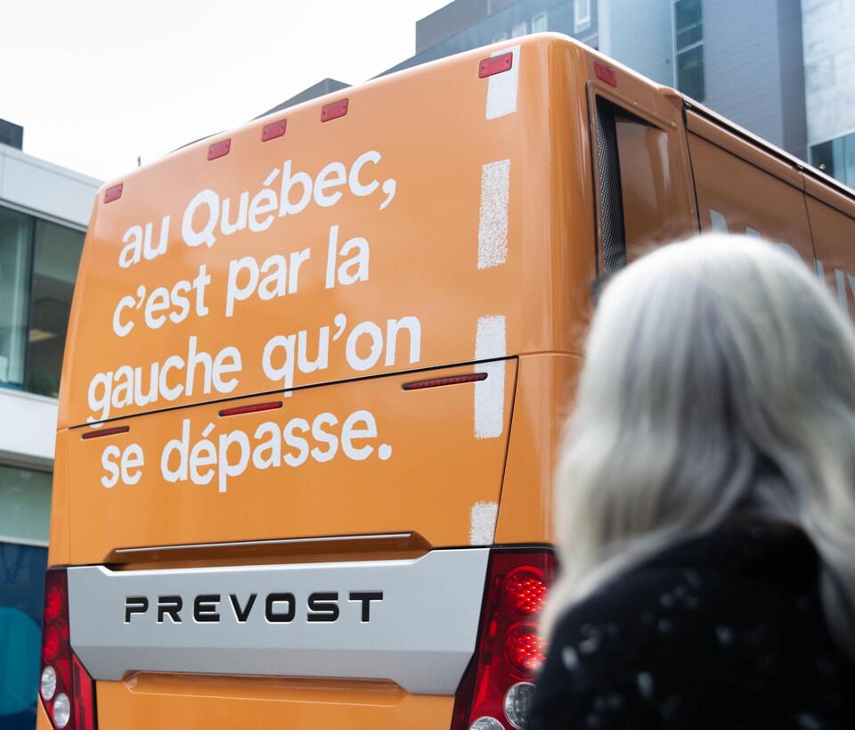 Un autobus orange. On y lit « Au Québec, c'est par la gauche qu'on se dépasse ». On y voit la co-porte-parole de Québec Solidaire, Manon Massé, de dos. 