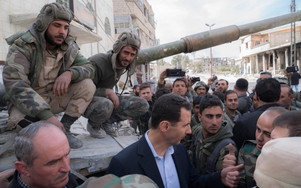 M. Assad en tenue décontractée, entouré par des soldats, près de chars militaires.