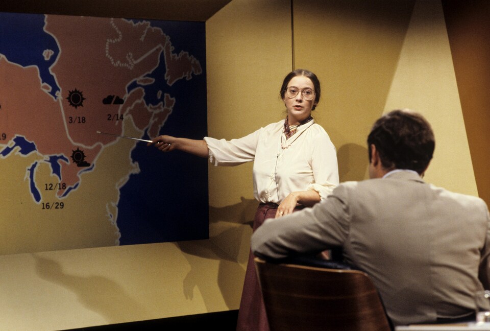 Dans un studio de télévision, la météorologue Jocelyne Blouin est debout, pointant une carte du Québec devant l'animateur et journaliste Bernard Derome, qu'on voit de dos, assis à un pupitre.