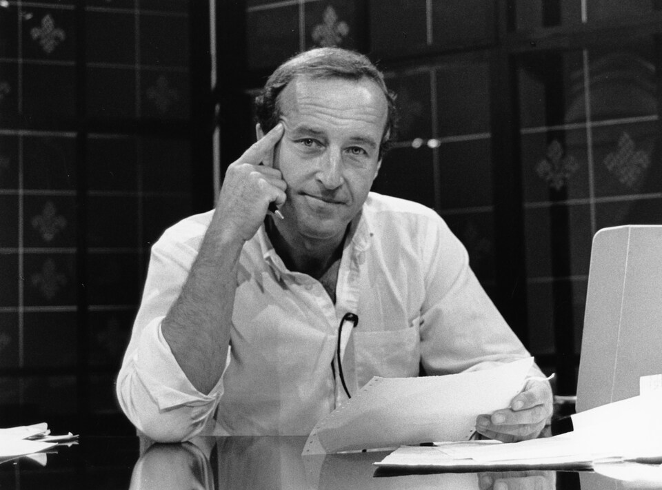 Dans un studio de télévision, l'animateur Bernard Derome, assis à une table, tient une feuille dans une main et le poing de son autre main, dont l'index est allongé, est appuyé sur sa tempe.