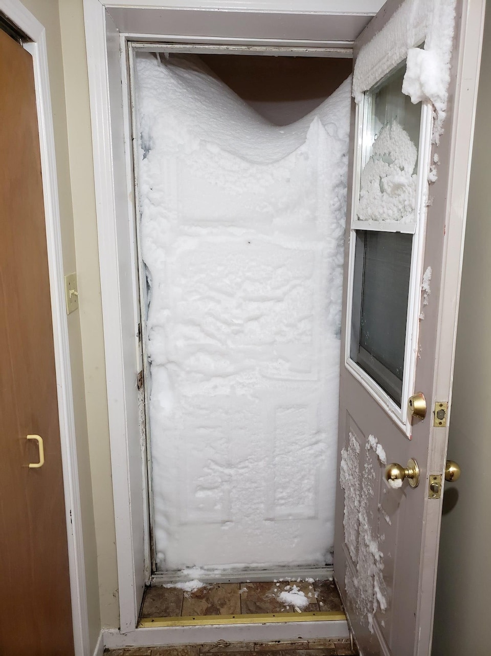 Une porte ouverte laisse voir un mur de neige qui bloque complètement l'accès et empêche de sortir d'une maison.