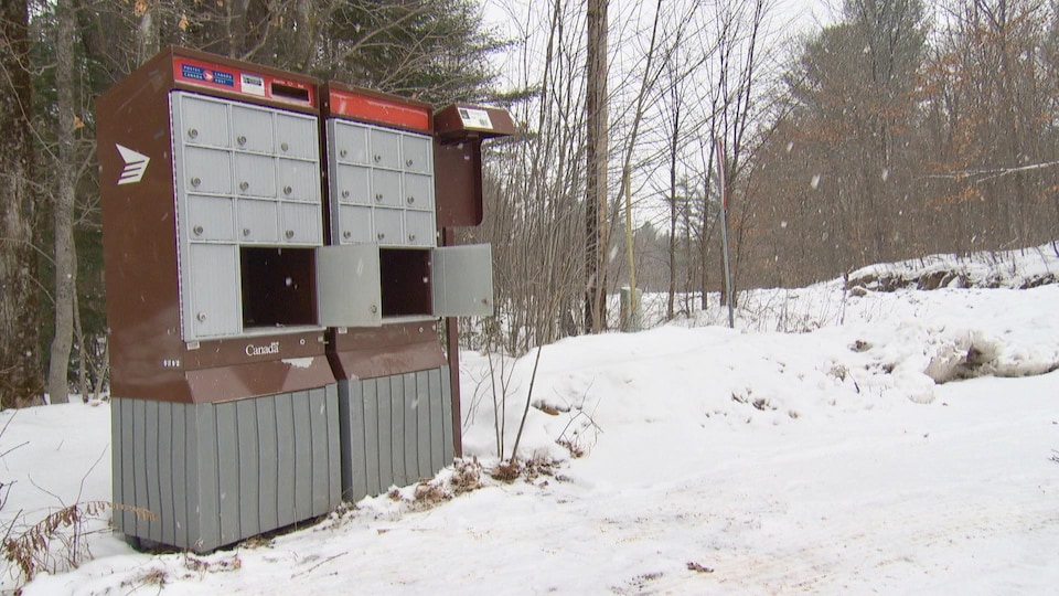 Une boîte aux lettres communautaire vandalisée, en bordure d'une route sous la neige. Deux cases sont complètement ouvertes. 