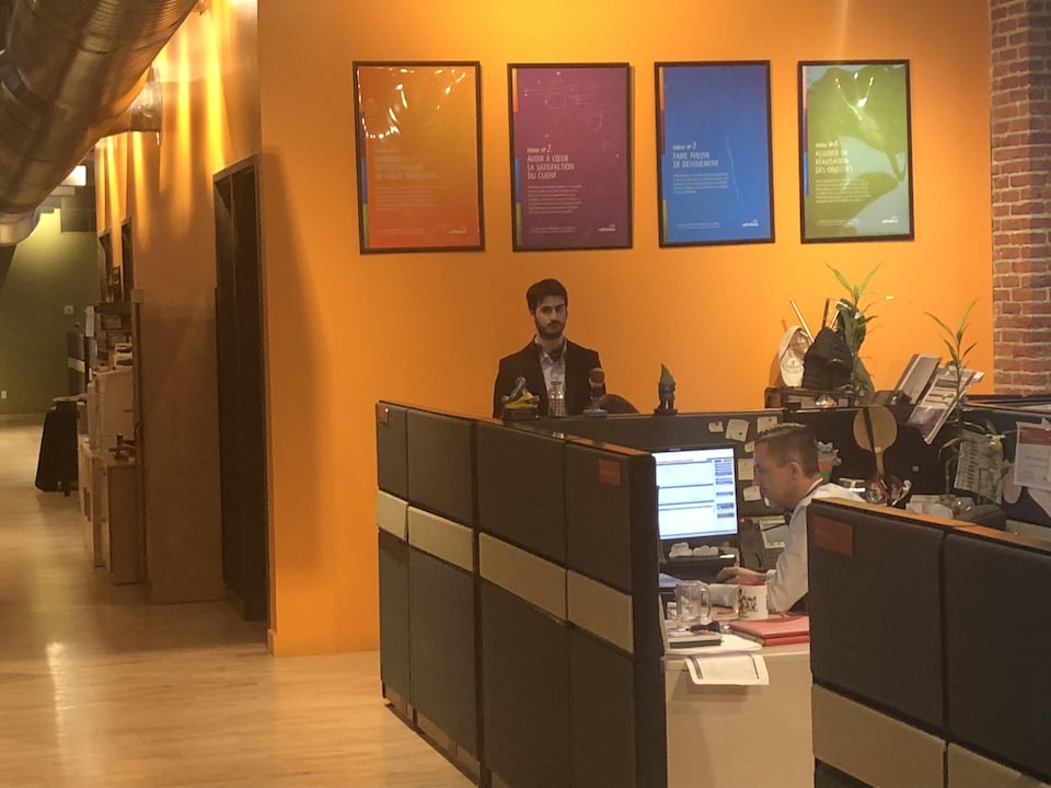 Un employé devant un ordinateur, et une personne debout. Au mur, des affiches en français.