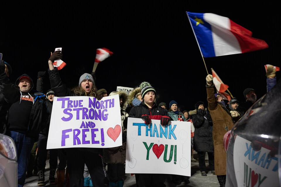 Des gens rassemblés dehors pendant la nuit. Ils agitent des drapeaux du Canada et un drapeau de l'Acadie et brandissent des pancartes en appui aux camionneurs.