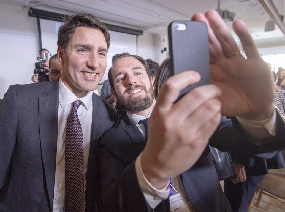 Le premier ministre Justin Trudeau posant pour un égoportrait avec un jeune homme barbu. Les deux hommes portent un veston-cravate.