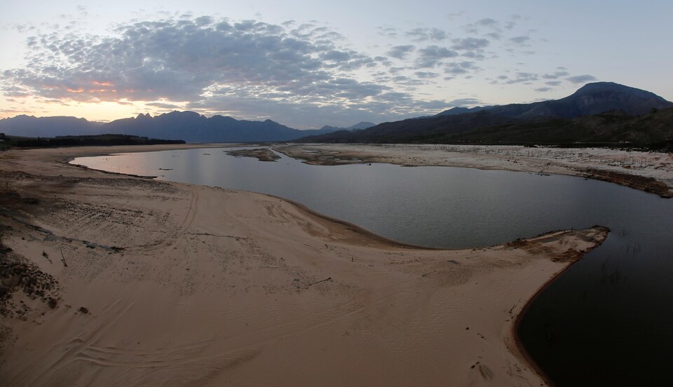 Le barrage Theewaterskloof est presque vidé de son eau, avec des montagnes sud-africaines au loin et le soleil se couchant à l'horizon.