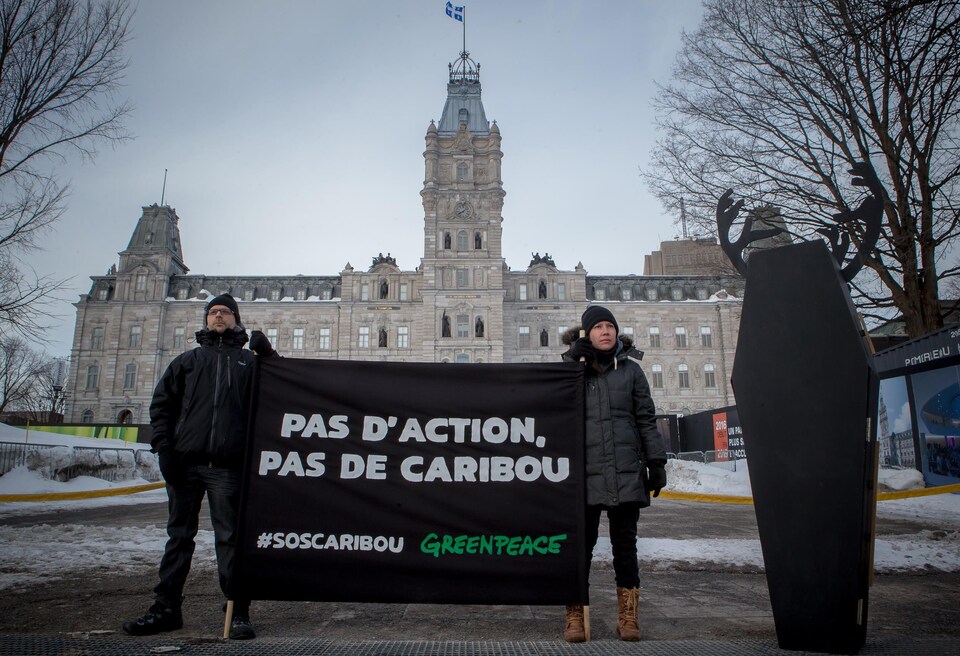 Deux personnes devant l'Assemblée nationale du Québec tiennent un drapeau avec le slogan « Pas d'action, pas de caribou » et le mot-clic « SOS caribous ».
