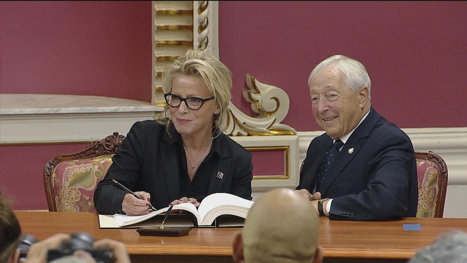 Caroline Proulx signe le registre officiel, assise à côté du lieutenant-gouverneur.