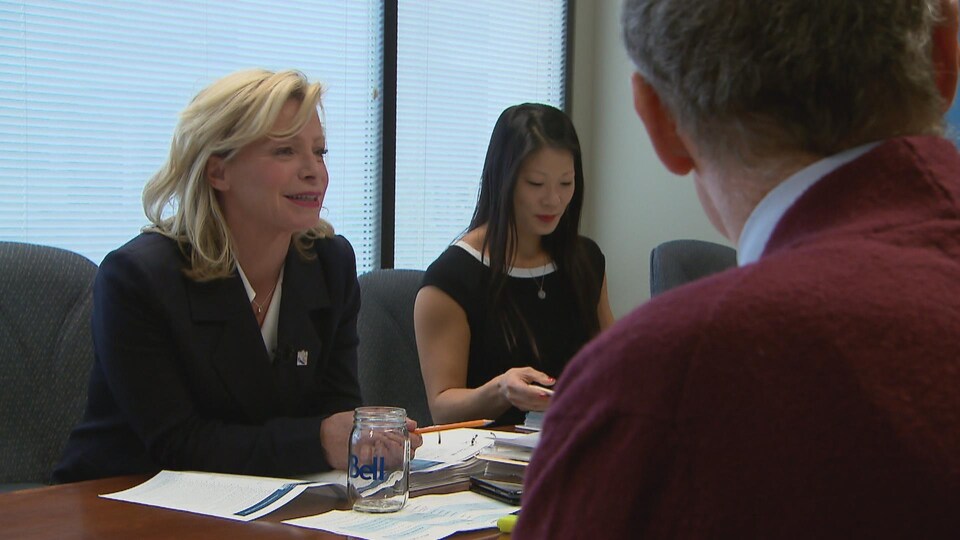 Caroline Proulx est assise à une table, à côté d'une femme et face à un homme.
