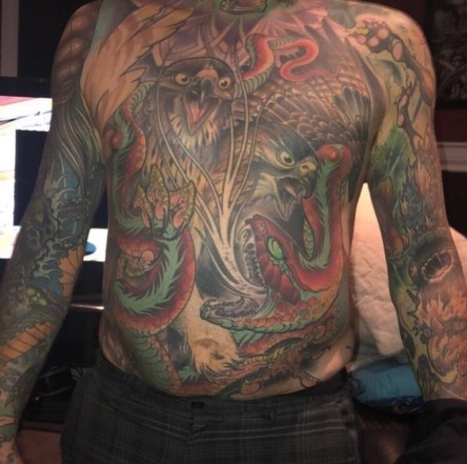 Le buste et les bras d'un homme torse nu, recouverts de tatouages. On y distingue deux faucons et un serpent stylisés.