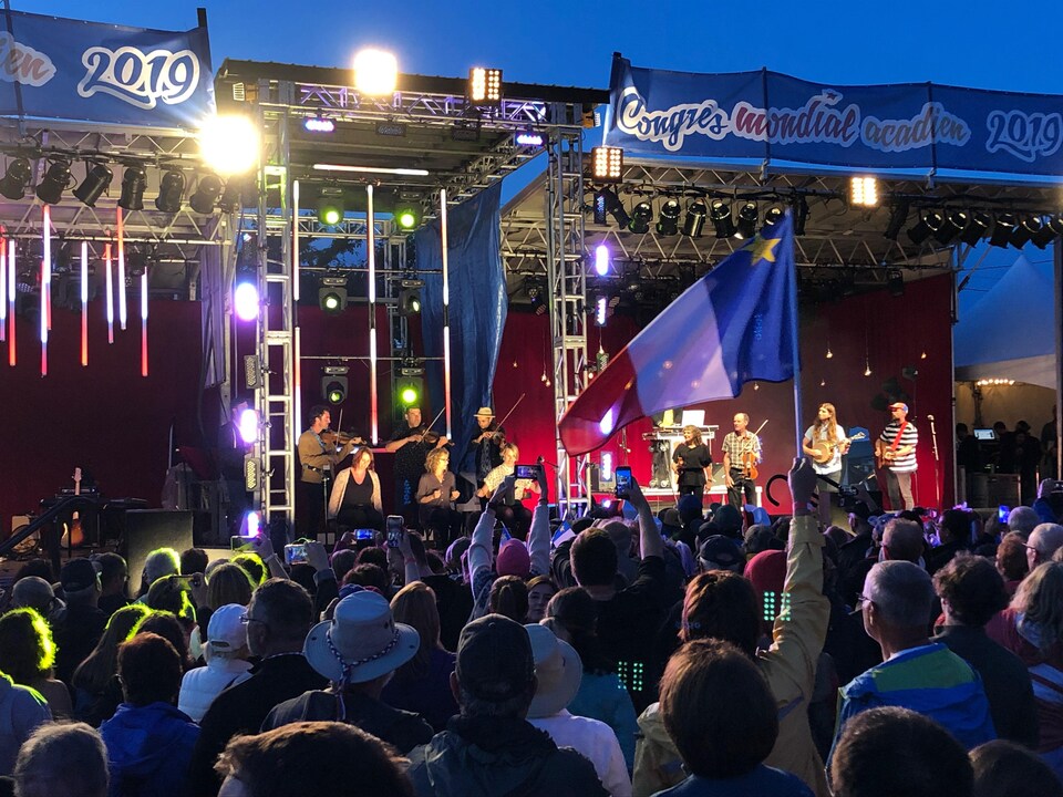 Quelqu'un dans la foule brandit un drapeau de l'Acadie alors qu'un groupe de musiciens est sur scène.