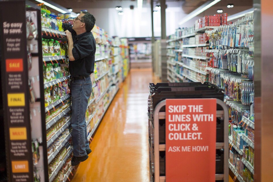 A man sits on the bottom shelf of a grocery display and struggles to grab an item from the top shelf.