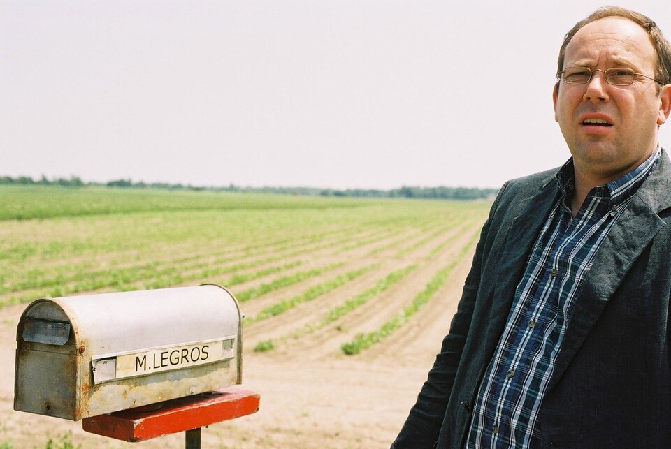 Une scène du film « Congorama » où l'on voit un acteur debout à côté d'une boîte aux lettres, devant un champ cultivé