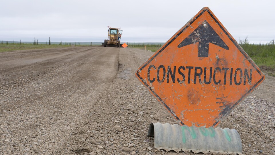 Un panneau de construction très usagé sur une route de gravier devant un camion de construction qui gratte la route au loin. 