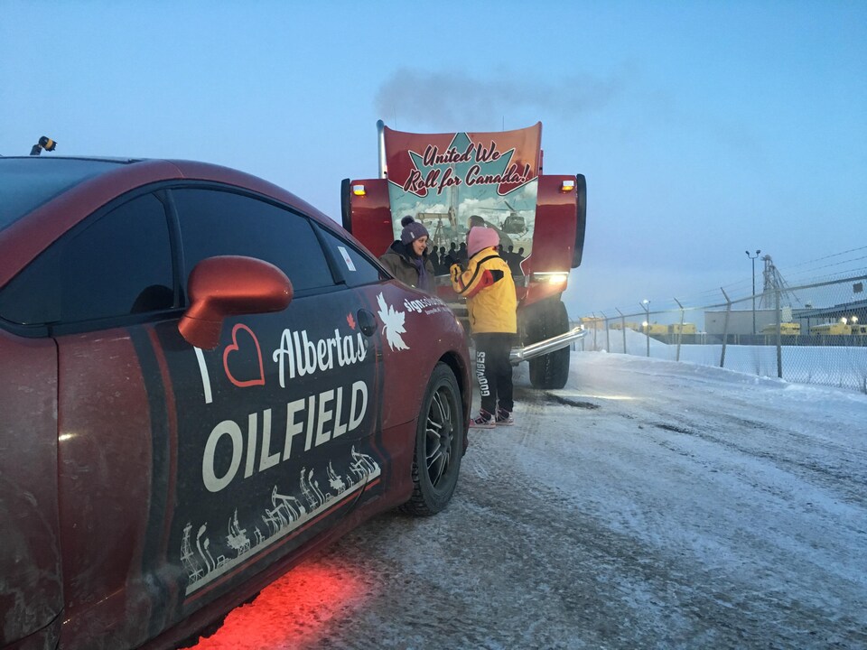 Une voiture rouge est en premier plan avec le message suivant sur la portière: « J'aime les gisements pétroliers de l'Alberta », écrit en anglais. En second plan se trouve un camion où il est écrit: « United we roll for Canada! ».
