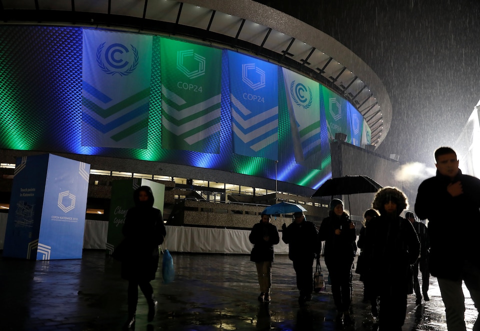 Des personnes marchent sous la pluie devant un stade orné de banderoles annonçant la COP24.