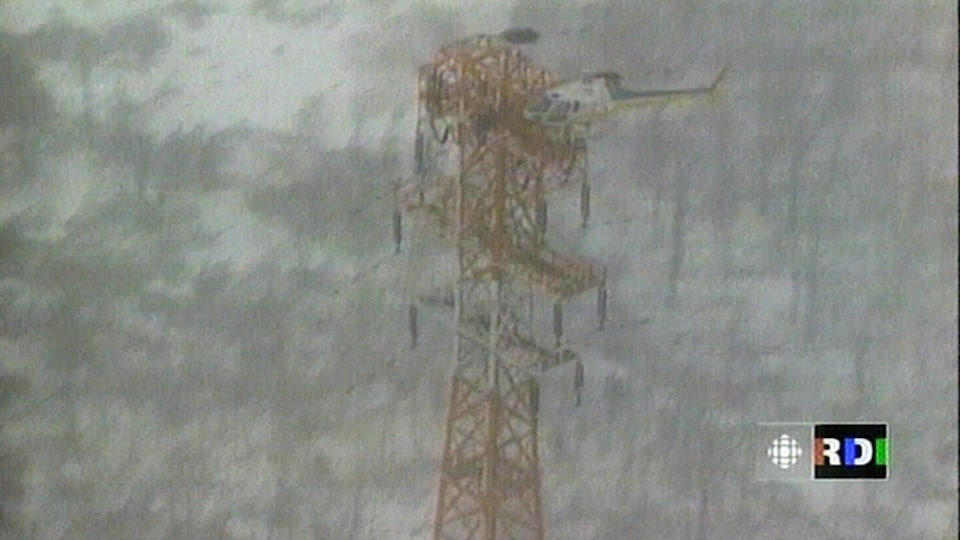 Un hélicoptère transporte deux monteurs de ligne d'Hydro-Québec au-dessus d'un immense pylône électrique.