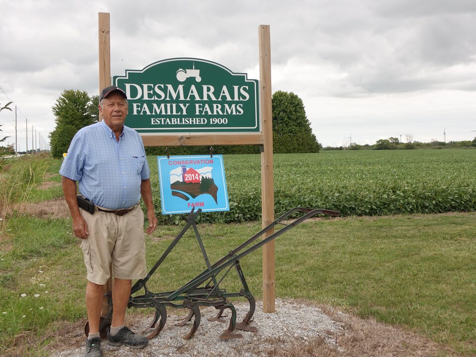 On voit M. Desmarais devant l'affiche où est écrit « Desmarais Family Famrs, Established 1900 ». En arrière-plan, un champ.