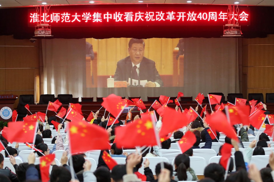 Des gens réunis devant un écran où est diffusé le discours du président chinois agitent des drapeaux à l'effigie de la Chine.