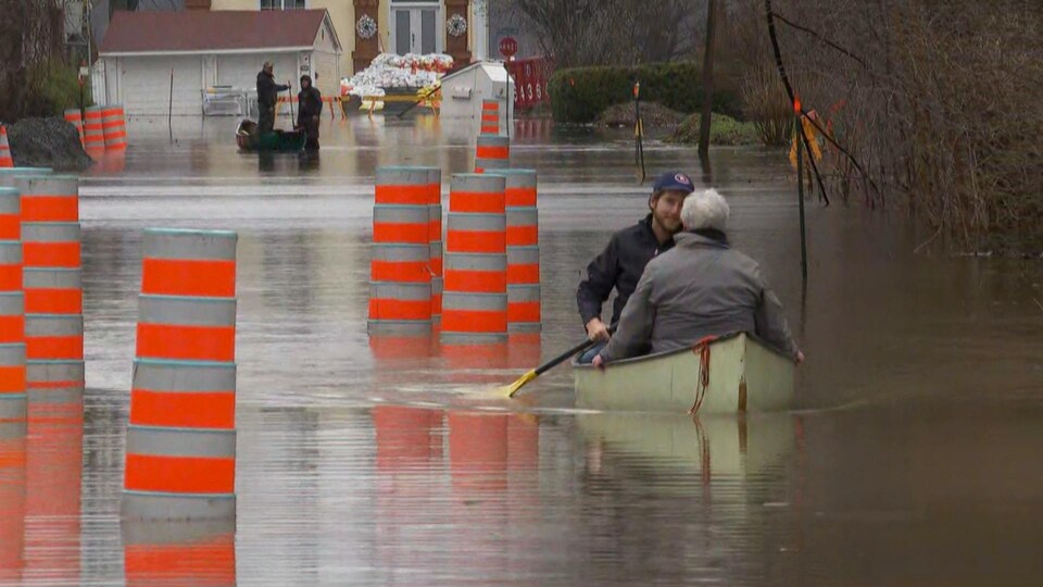 Deux personnes sont dans une chaloupe dans la rue inondée, contournant des cônes orange.